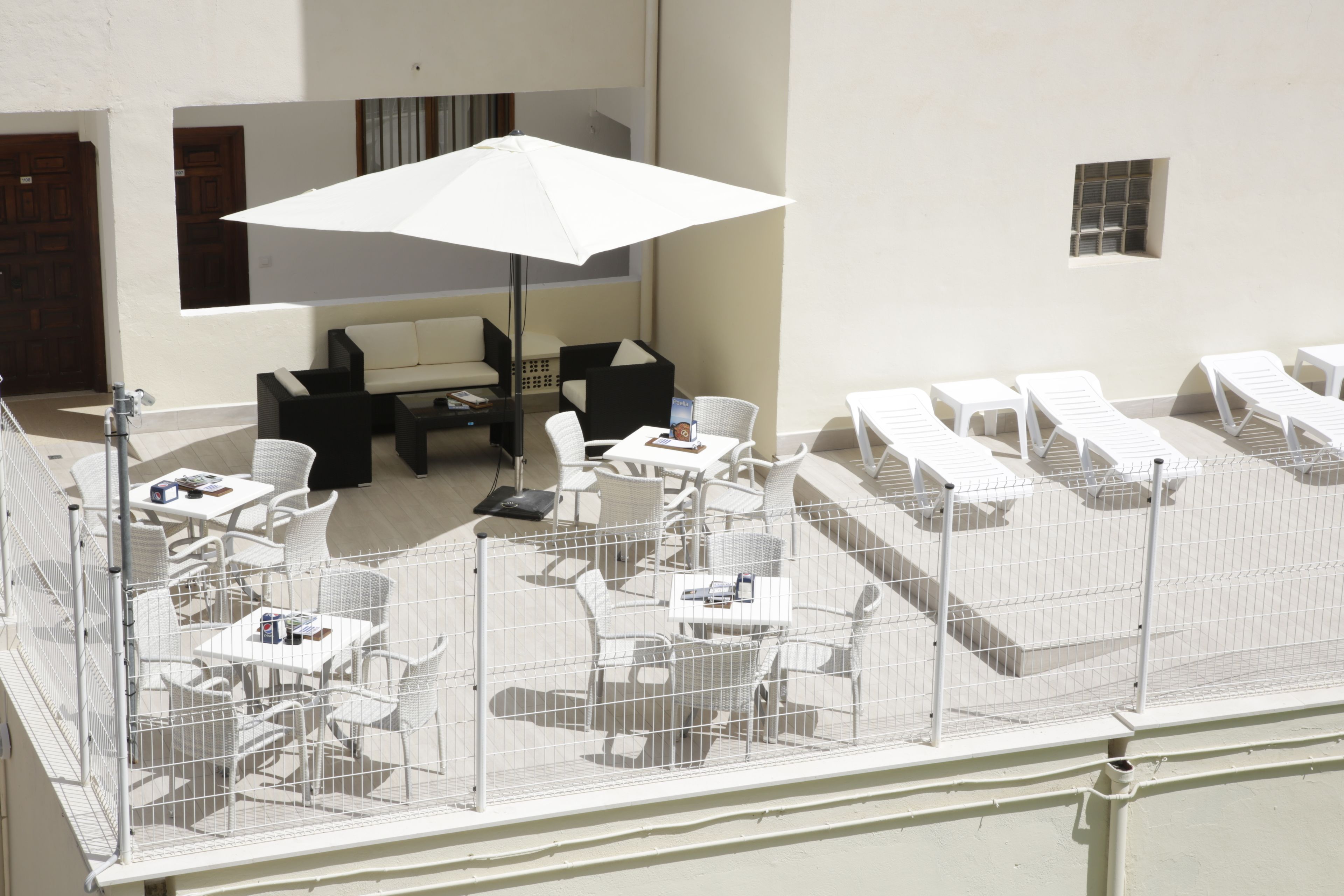 Een terras op het dak met witte loungestoelen en tafels, een witte parasol, en een omheind gebied met extra zitplaatsen.
