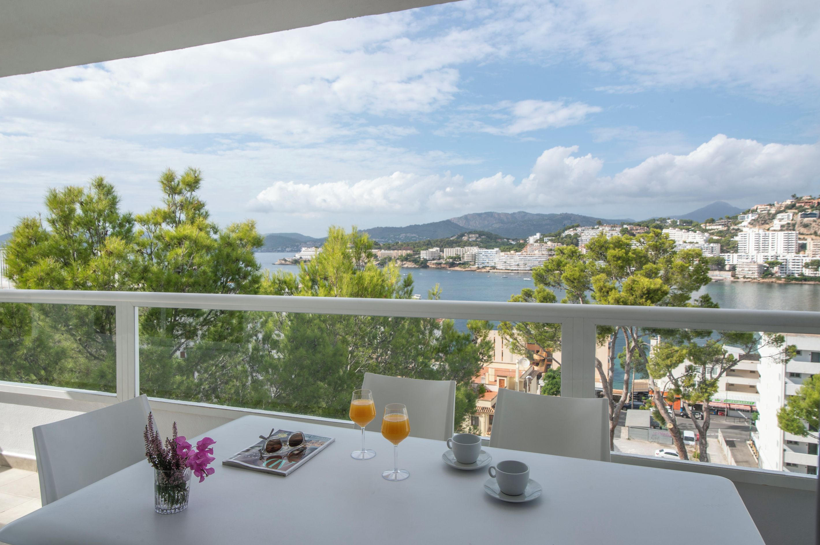 A balcony with a table set for two, overlooking a coastal cityscape with buildings and a body of water.