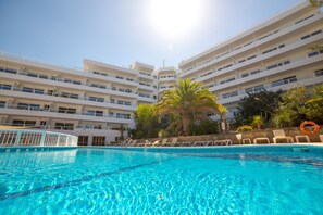 A swimming pool with clear blue water, surrounded by a white multi-story building with balconies, palm trees, and lounge chairs. - Pierre & Vacances Mallorca Portofino (Calvia)