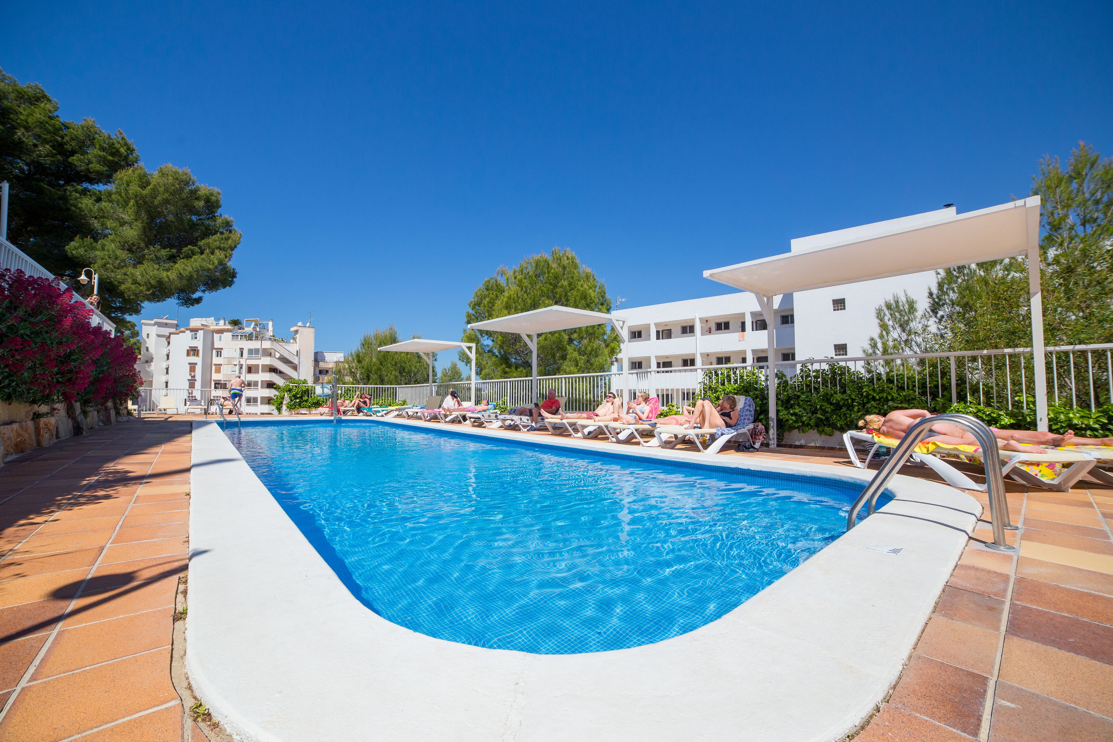 A swimming pool with sun loungers and umbrellas, surrounded by buildings and trees.