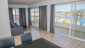 A modern living room with a sofa, a dining table, and chairs. Large windows offer a view of a body of water and buildings. - Pierre & Vacances Mallorca Portofino (Calvia)