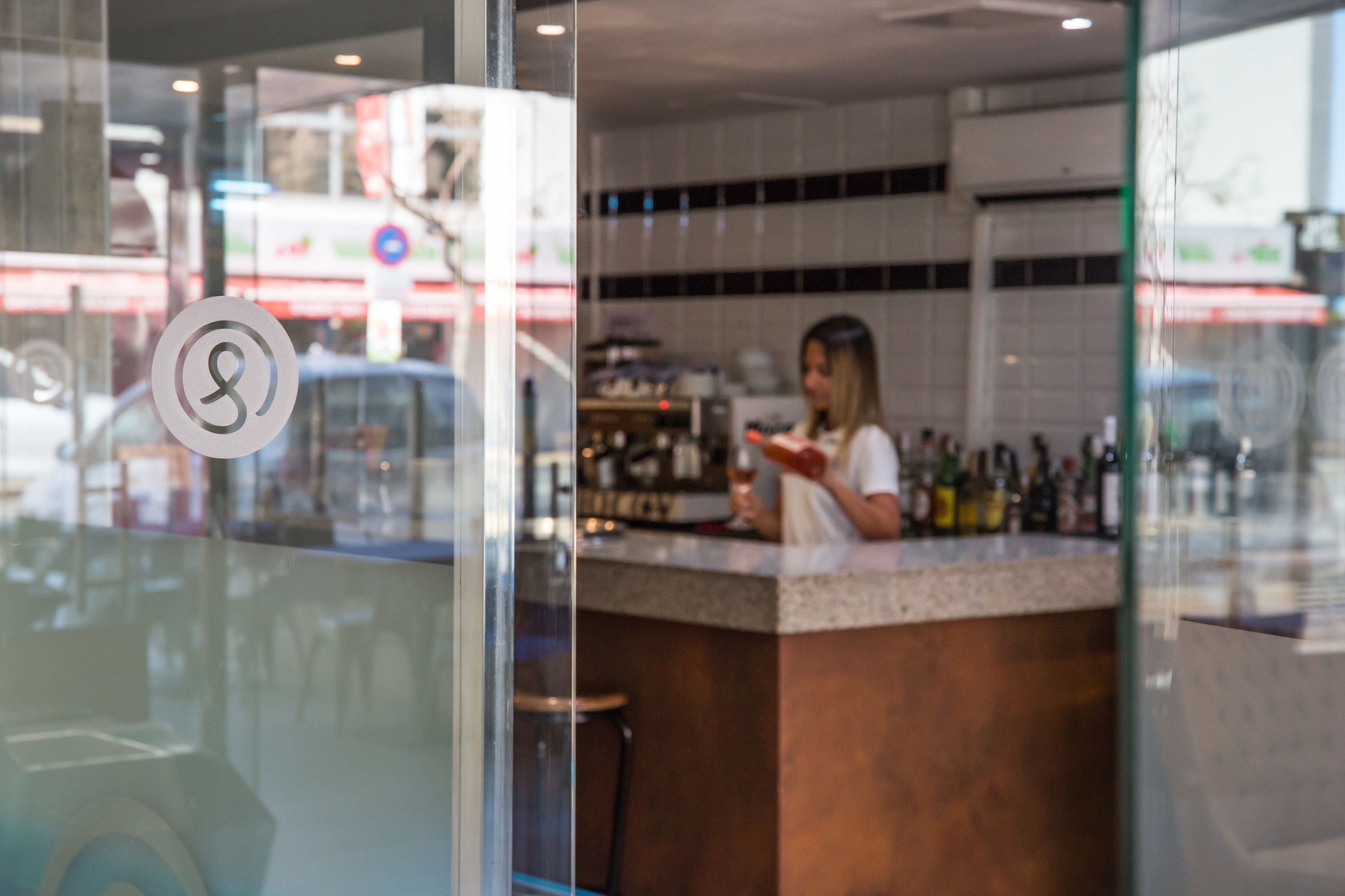 Un espace bar avec un comptoir et des tabourets, une personne debout derrière le comptoir, et une cloison en verre ornée d’un logo.