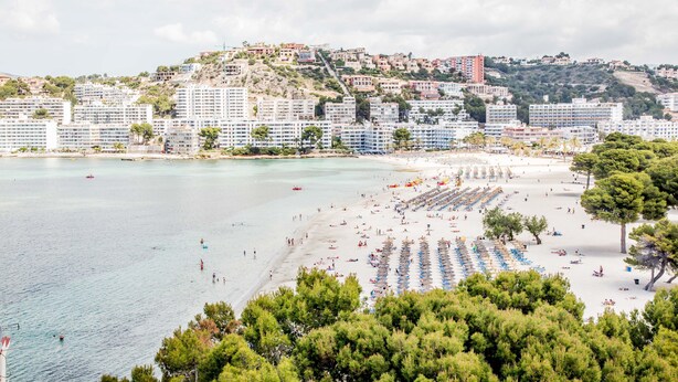 A coastal cityscape with a beach, rows of beach umbrellas, and buildings along the hillside. - Pierre & Vacances Mallorca Portofino (Calvia)