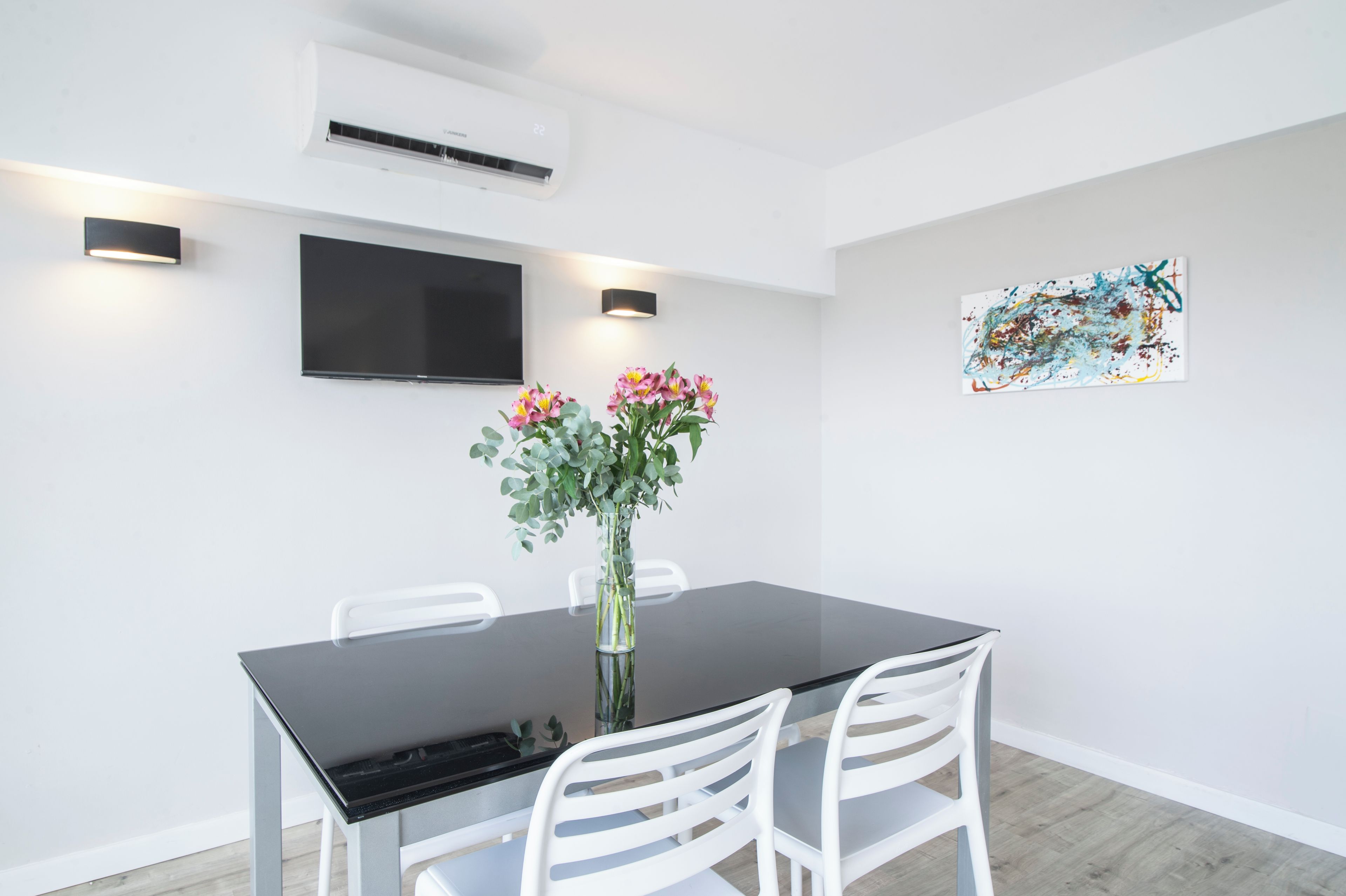 A modern dining area with a black table, white chairs, a vase with flowers, a flat-screen TV, and a wall-mounted air conditioner.