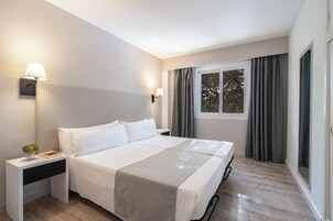 Habitación de hotel con cama doble, lámparas de noche, mesita de noche, ventana con cortinas y vistas a árboles.