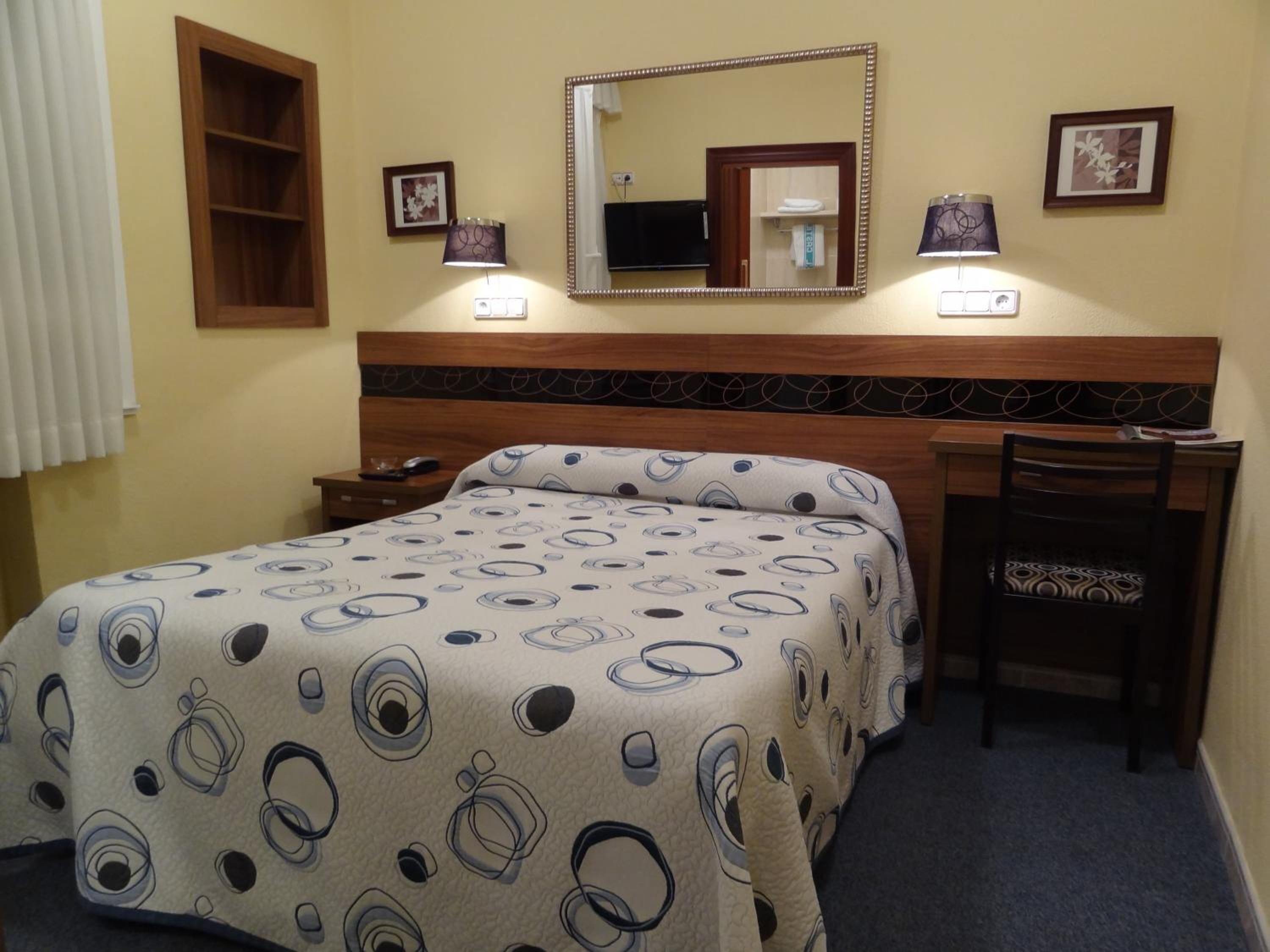 Una habitació d'hotel amb un llit, una taula de treball, una cadira, un mirall i dues làmpades penjades a la paret.