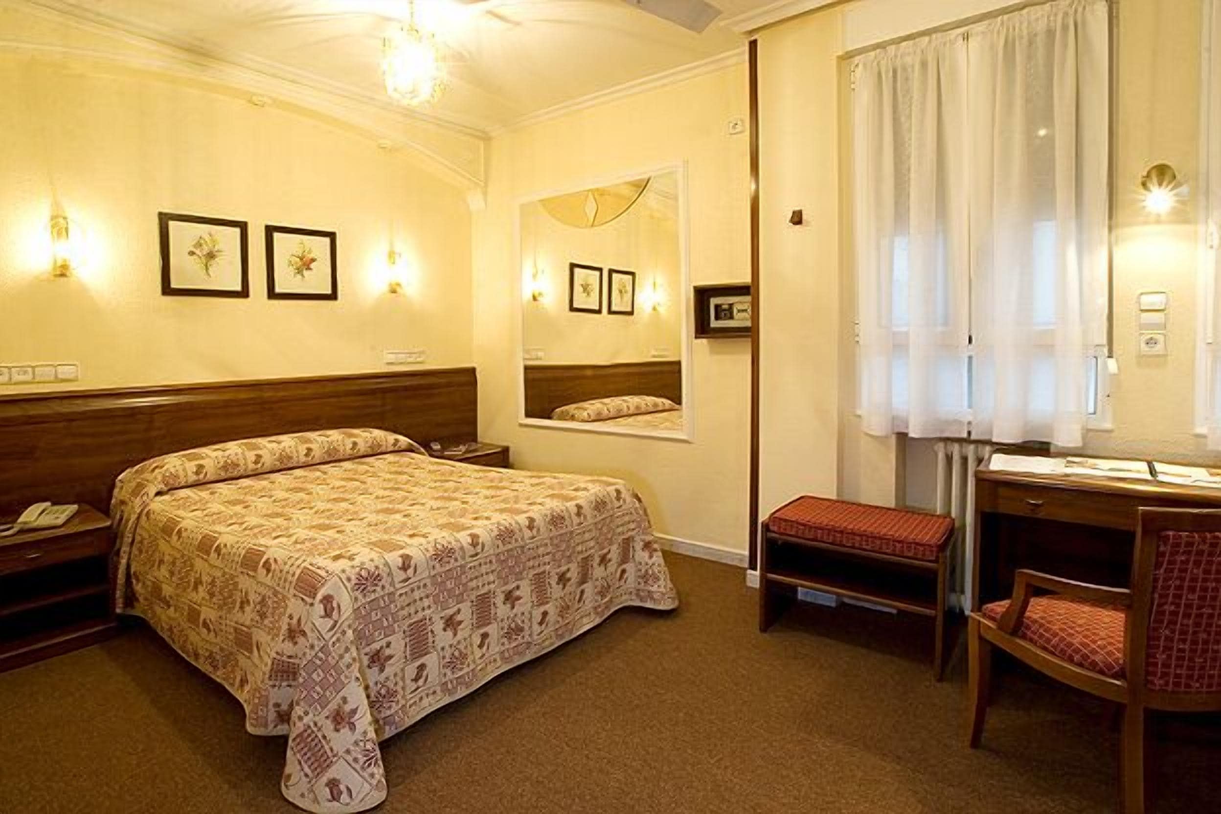 Ein Hotelzimmer mit Bett, Schreibtisch, Stuhl, kleinem Tisch, Bank, Spiegel und Wandleuchten.