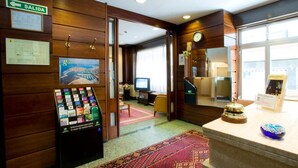 Vestíbulo de hotel con mostrador de recepción, panel informativo digital, reloj y ventana con vistas a la ciudad.