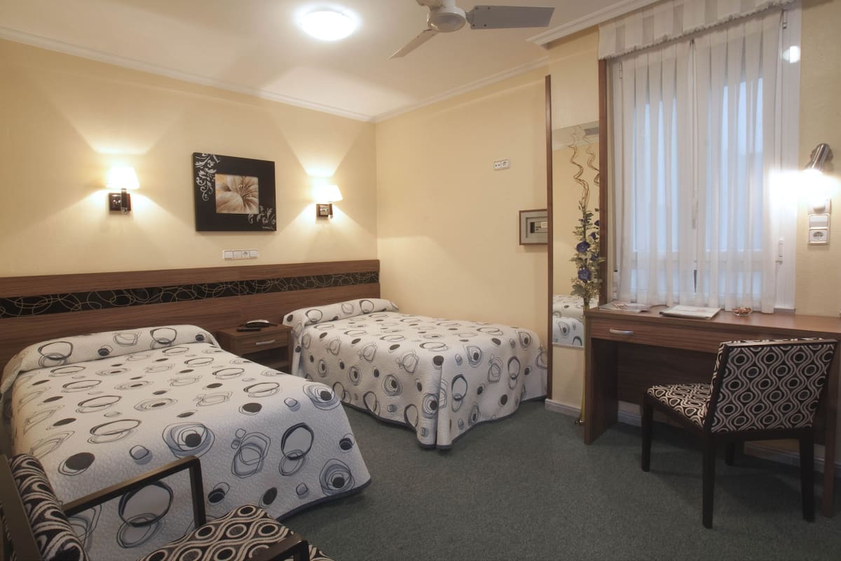 Habitació d'hotel amb dos llits, una escriptori, una cadira, un ventilador de sostre i una finestra amb cortines.