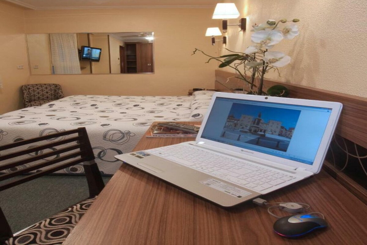 Una habitación de hotel con una cama, un escritorio con una computadora portátil, una silla, una lámpara de escritorio y una orquídea en maceta.