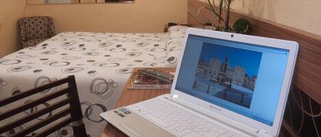 A hotel room with a bed, a desk with a laptop, a chair, a desk lamp, and a wall-mounted TV.
