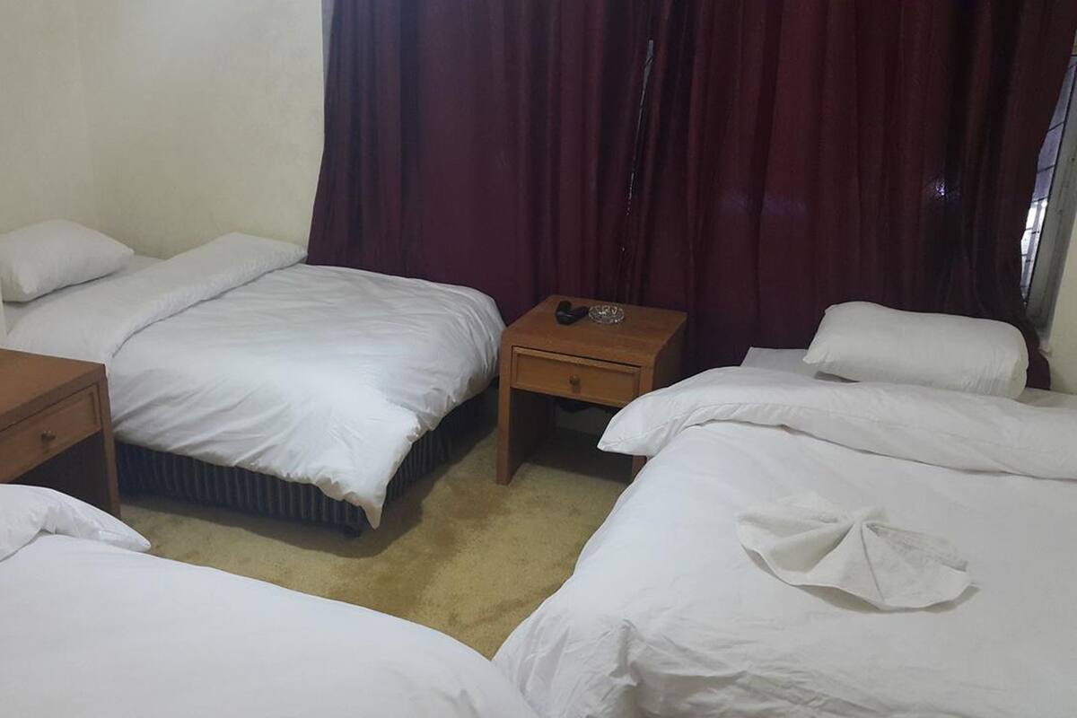 Une chambre d’hôtel avec deux lits simples, une table de chevet en bois, une petite table et une fenêtre avec des rideaux foncés.
