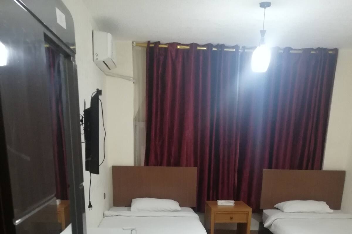 Une chambre d’hôtel avec deux lits simples, un bureau et une petite table. Il y a un radiateur au mur et une poubelle près de la porte.