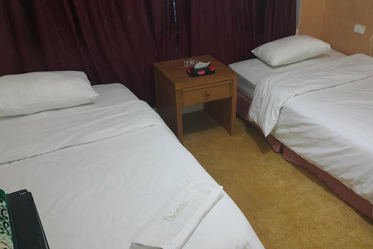 Une chambre d’hôtel avec deux lits simples, une table de chevet en bois sur laquelle se trouve une boîte de mouchoirs, et un sol recouvert d’un simple tapis.