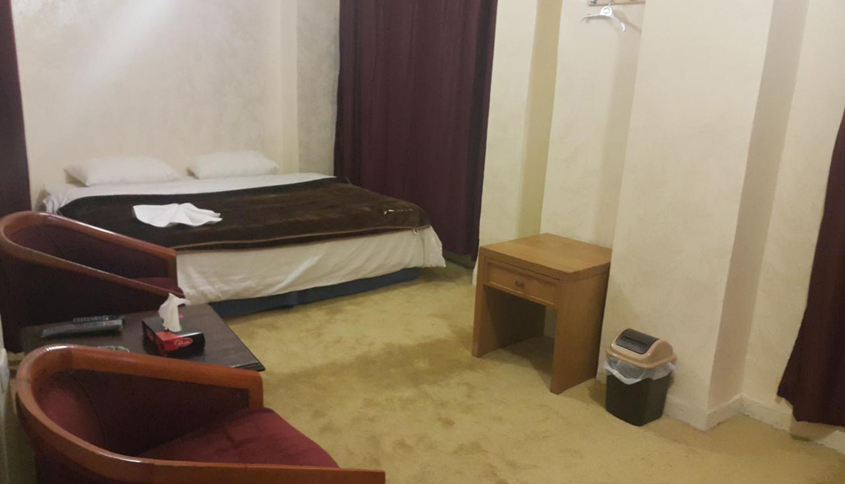 Una habitación de hotel con una cama, una mesita de noche de madera, un cesto de basura y una silla.