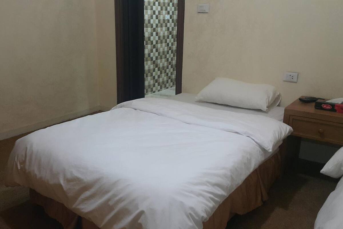 Un lit simple avec du linge de lit blanc, dans une pièce aux murs de couleur beige et dont le mur derrière le lit est orné d’un motif à carreaux.