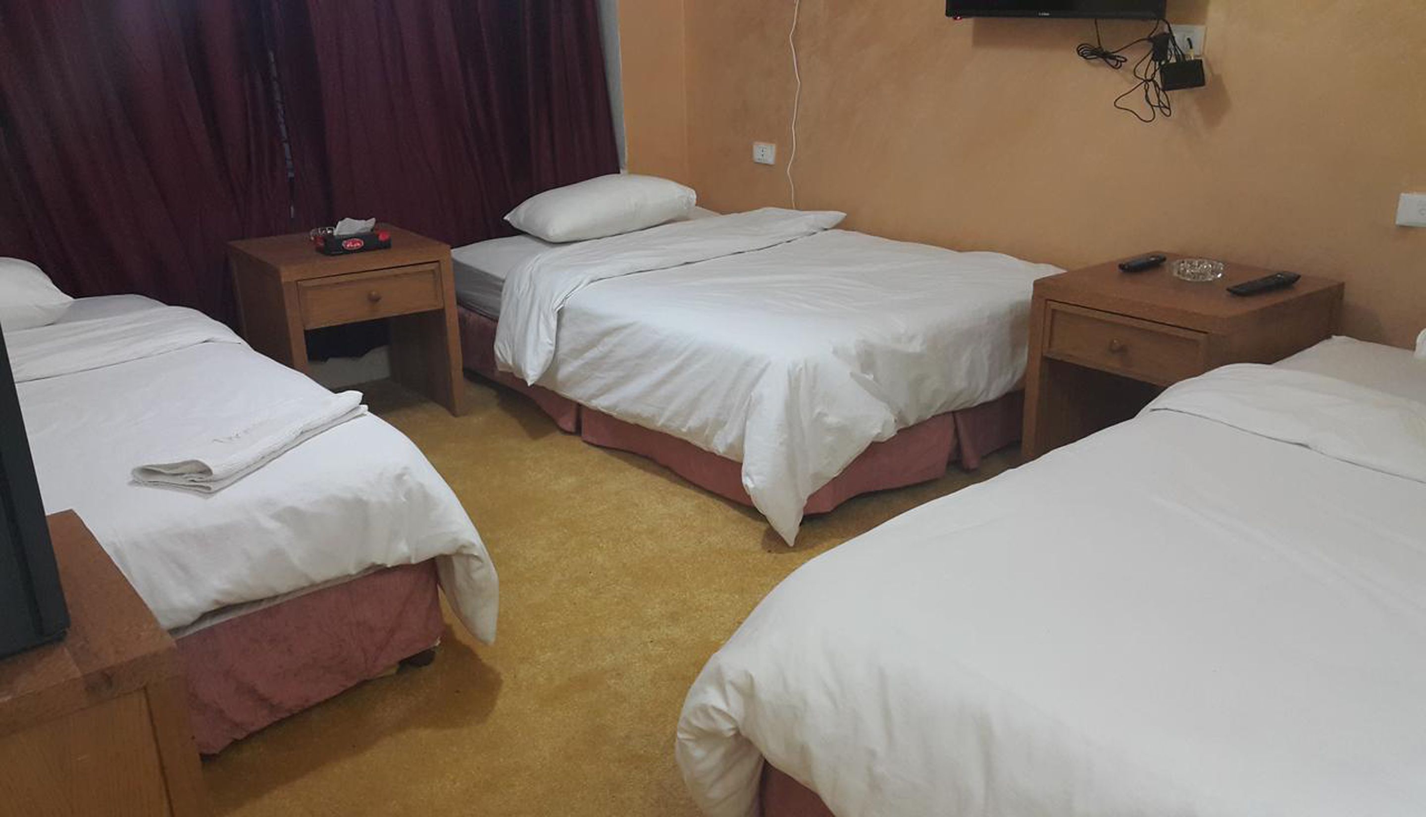 Quarto de hotel com três camas de solteiro, mesas de cabeceira de madeira, uma televisão de tela plana e uma pequena mesa com um controle remoto.