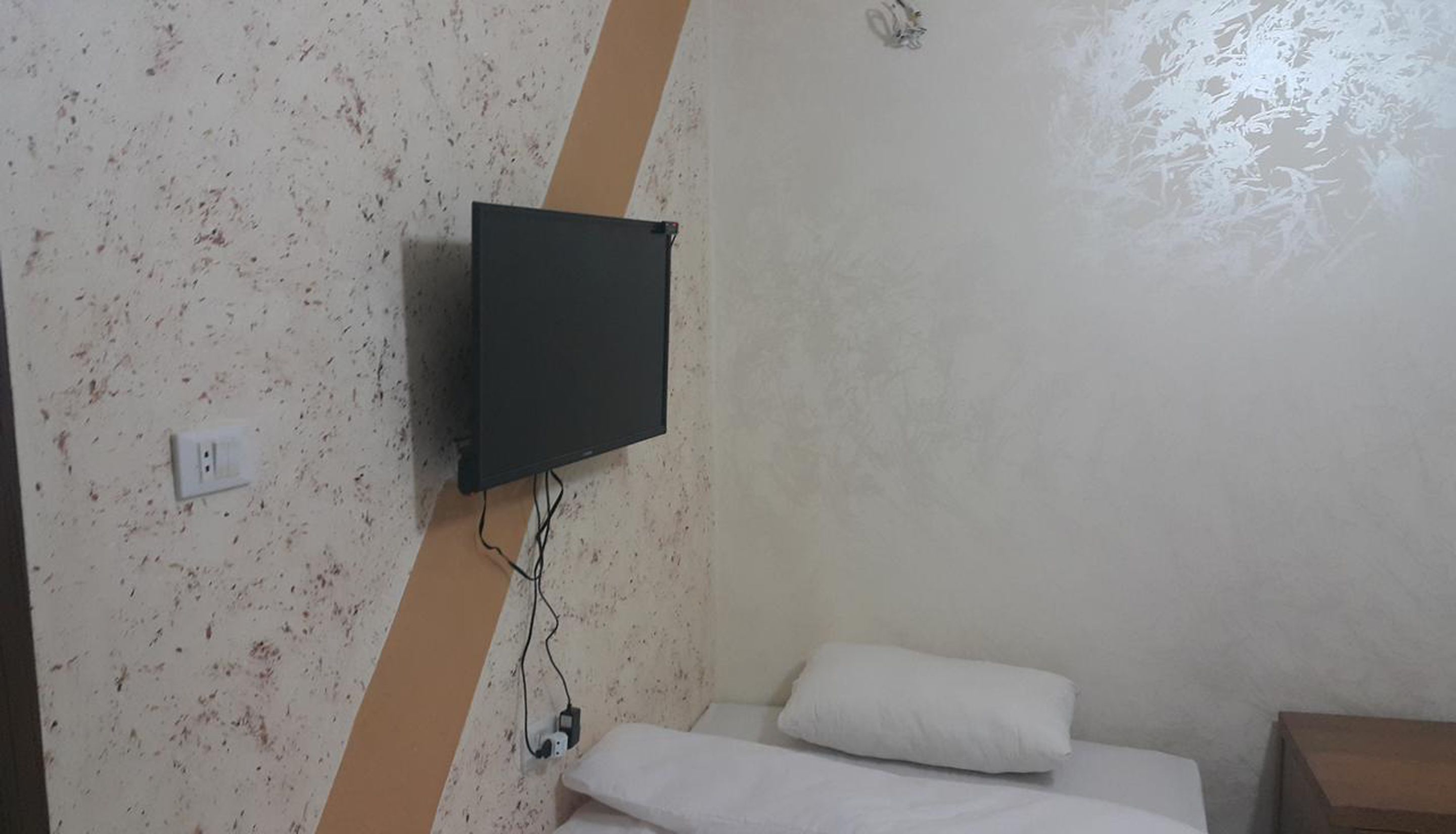 A room with a bed, a white pillow, a TV mounted on the wall, and a power outlet.