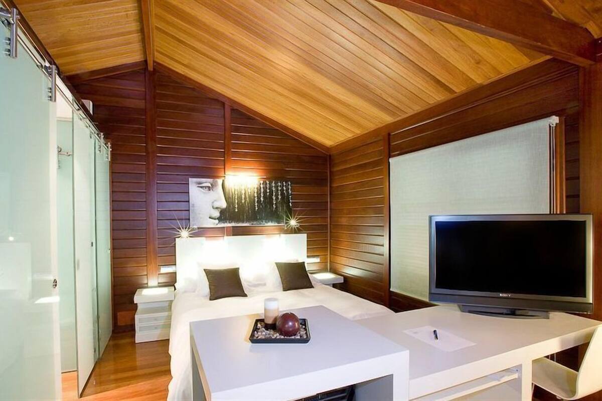 A modern bedroom with a wooden ceiling, a bed with white bedding, a bedside table with a TV, and a glass wardrobe.