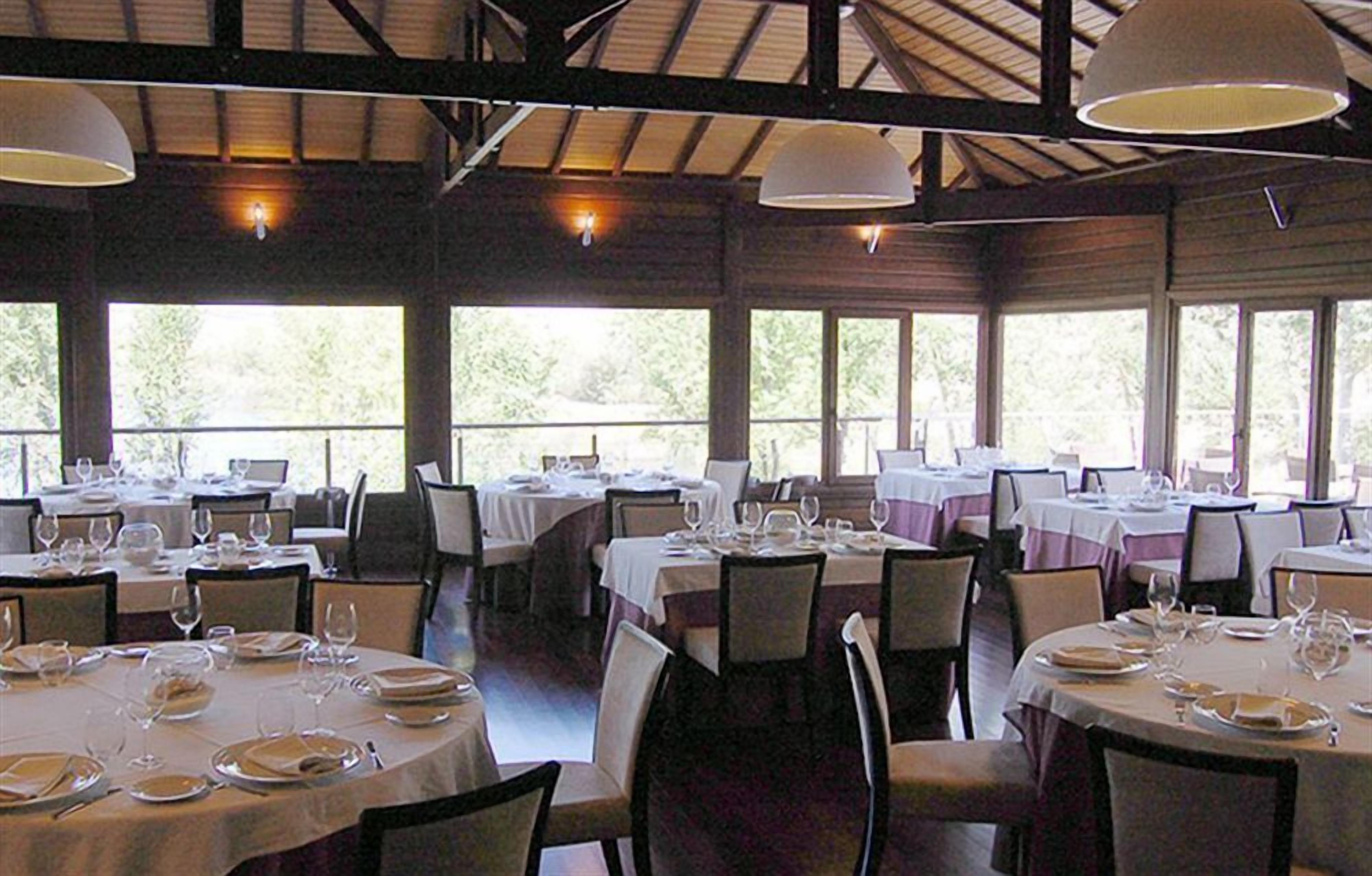 Un restaurante con mesas redondas dispuestas para comer, ventanas amplias y un techo alto con vigas a la vista.