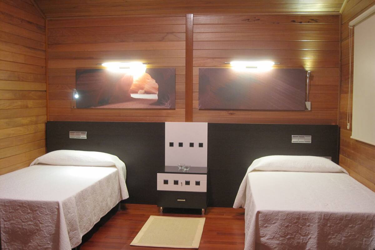 A room with two beds, wooden walls, a small table, and two wall-mounted TVs.
