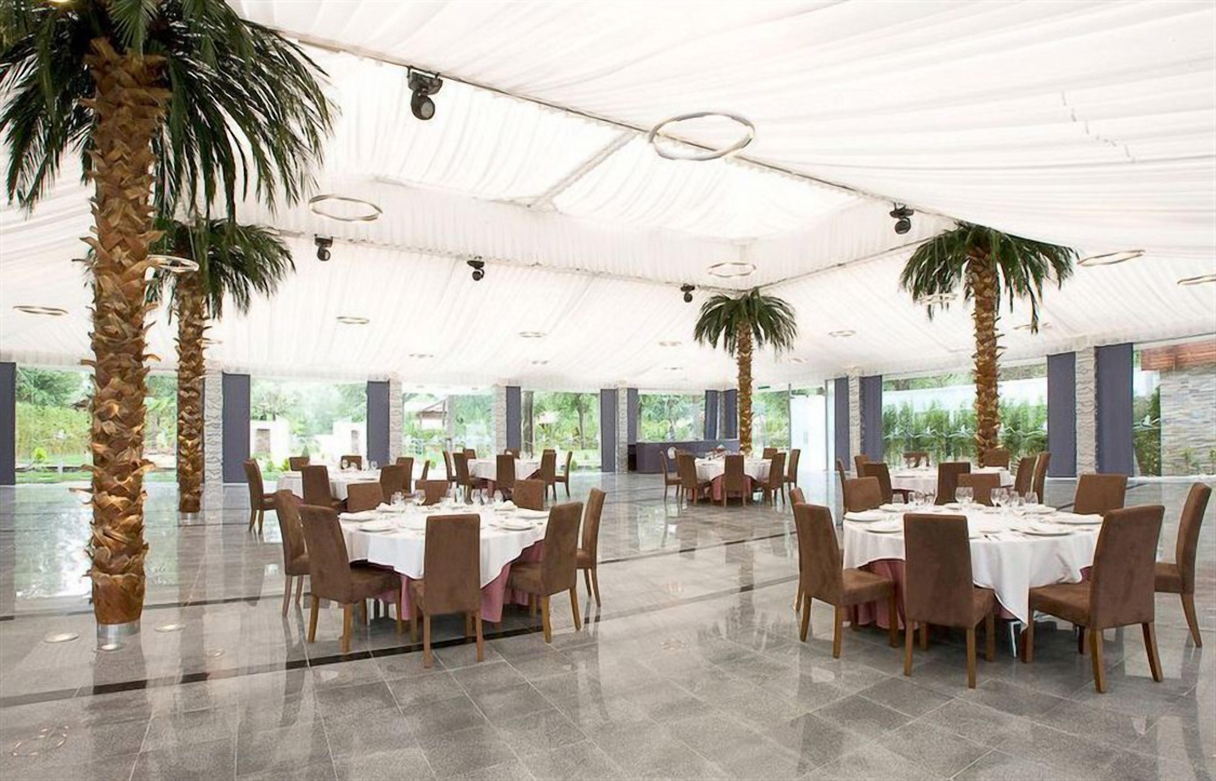 A spacious indoor dining area with round tables and chairs, large windows, and palm trees.