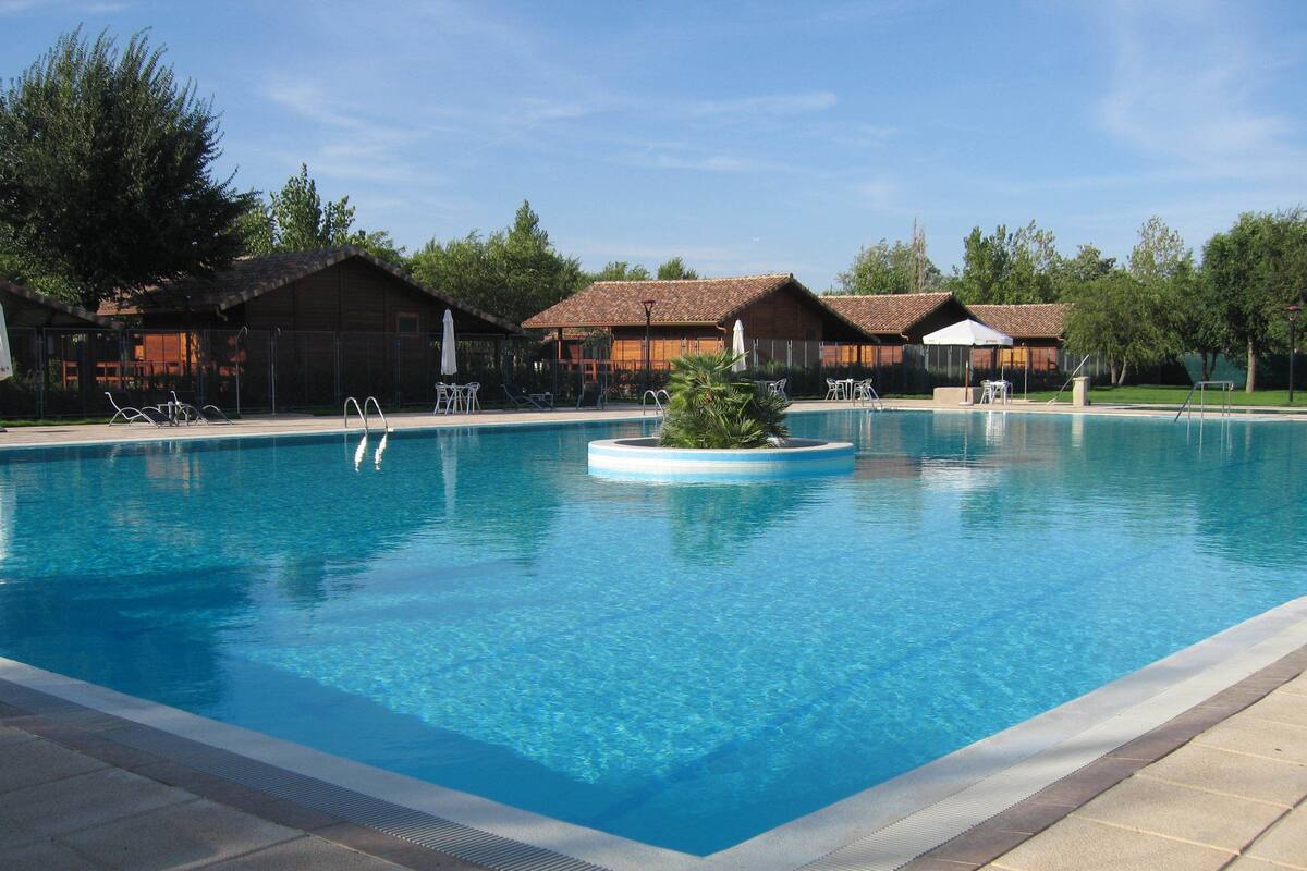 A rectangular swimming pool with clear blue water, surrounded by lounge chairs and umbrellas, and adjacent to a building with a tiled roof.