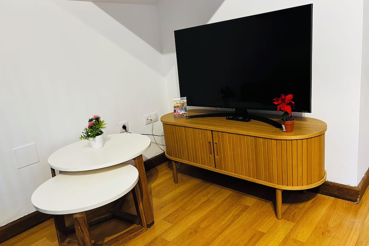 A small living space with a TV on a wooden cabinet, a white round table with two chairs, and a potted plant.