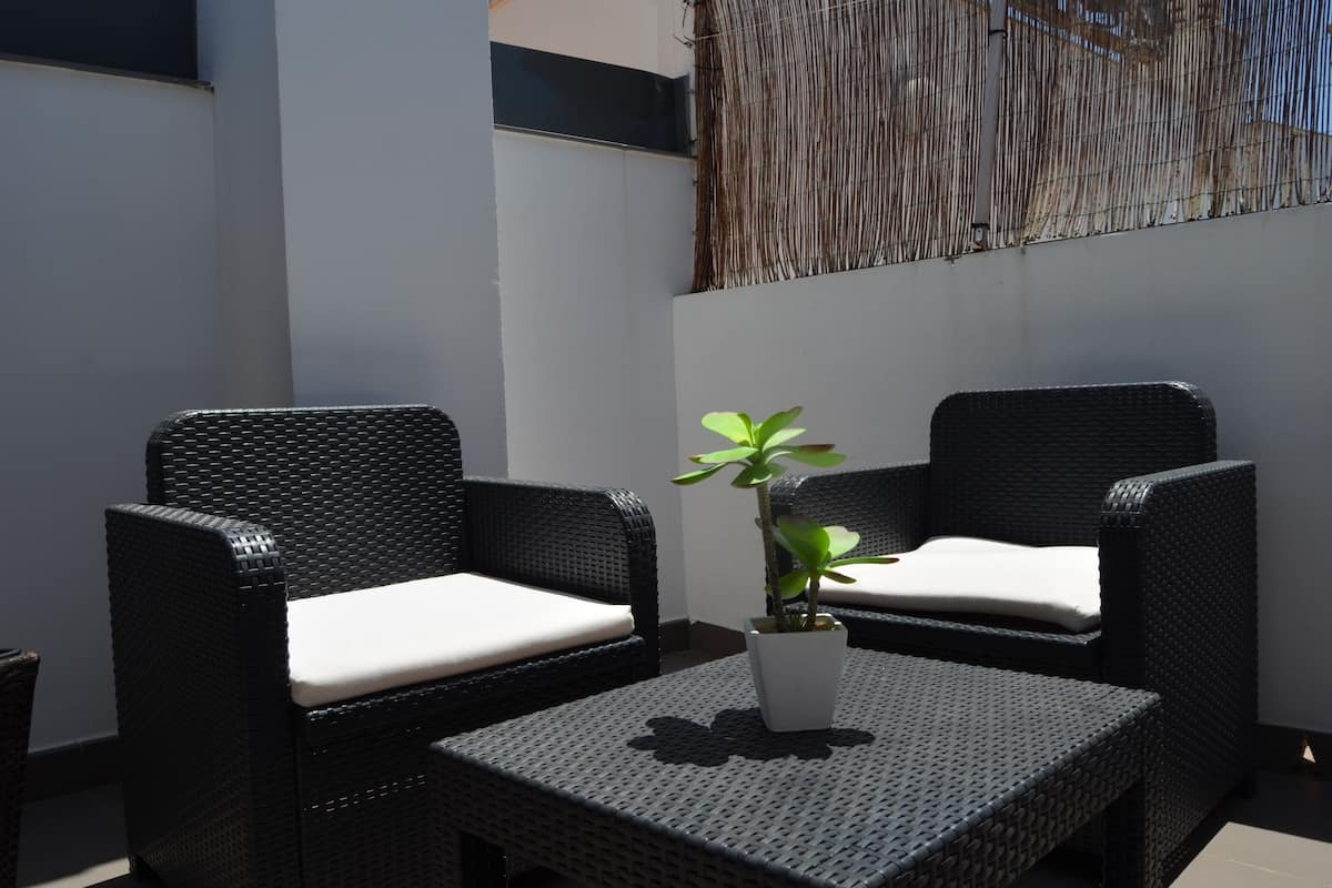 A small outdoor seating area with two wicker chairs and a table, a potted plant, and a woven wall decoration.