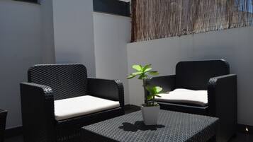 Un pequeño espacio al aire libre con dos sillas de mimbre, una mesa, una planta en maceta y un adorno de pared tejido.
