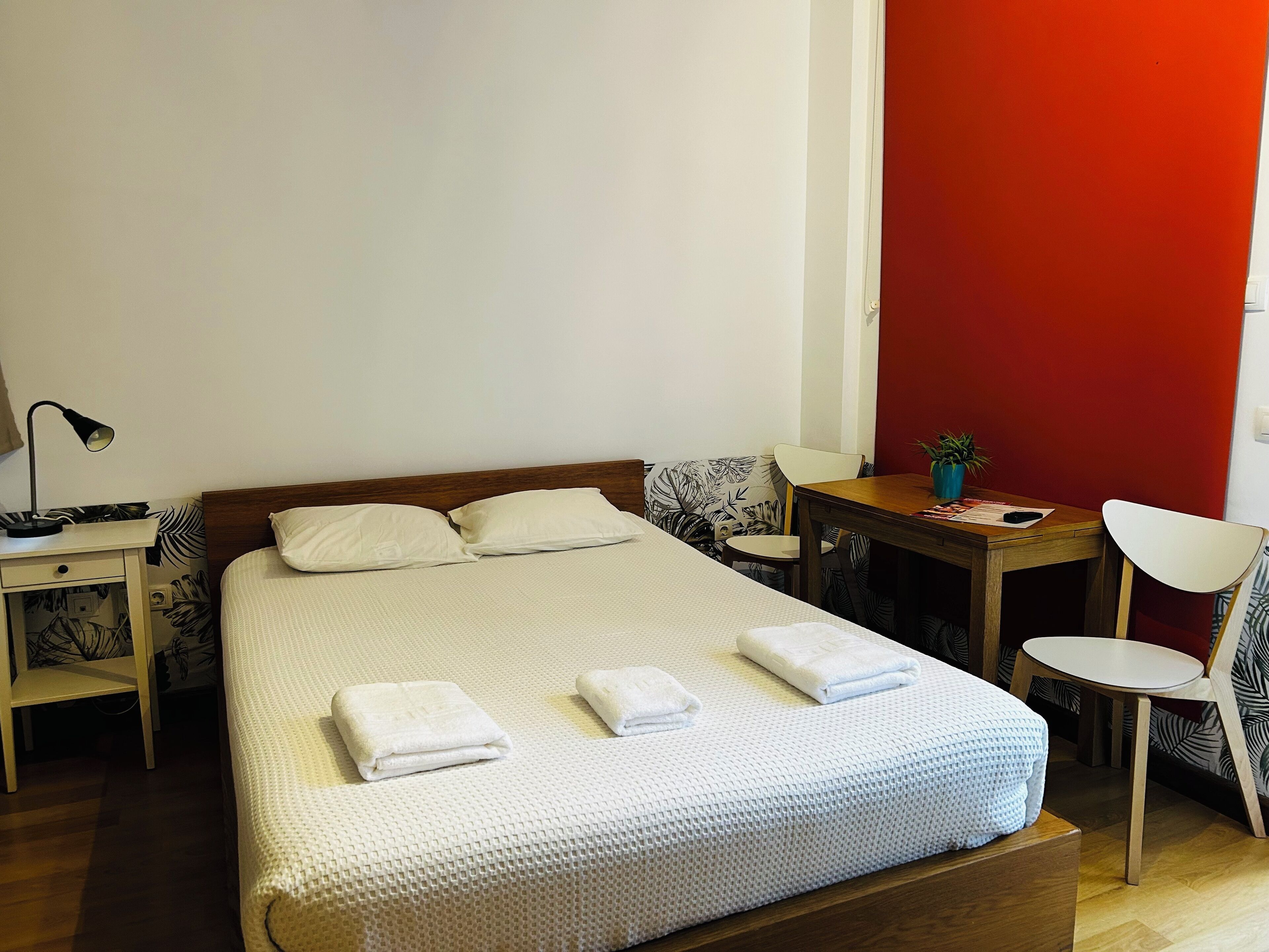 A neatly made bed with white linens and towels, a bedside table with a lamp, a small round table with a plant, and a red accent wall.