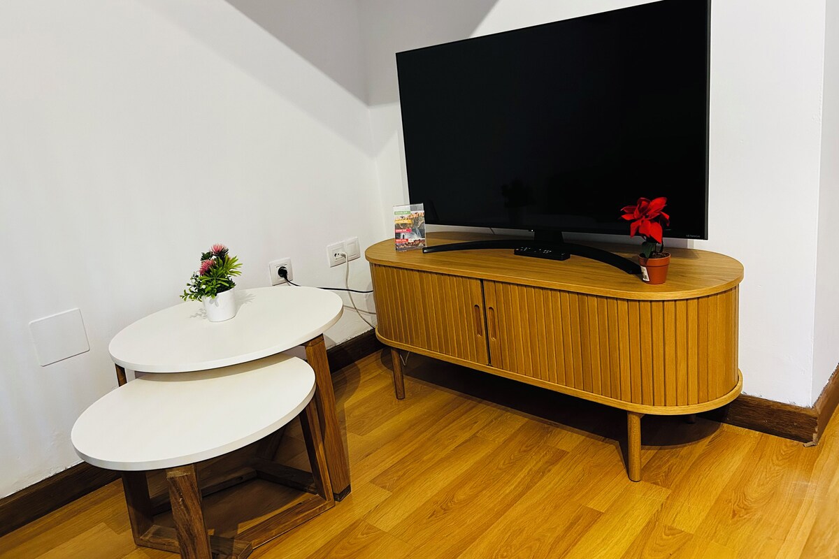 Een kleine leefruimte met een televisie op een houten kast, een witte ronde tafel met twee stoelen, en een plant in een pot.
