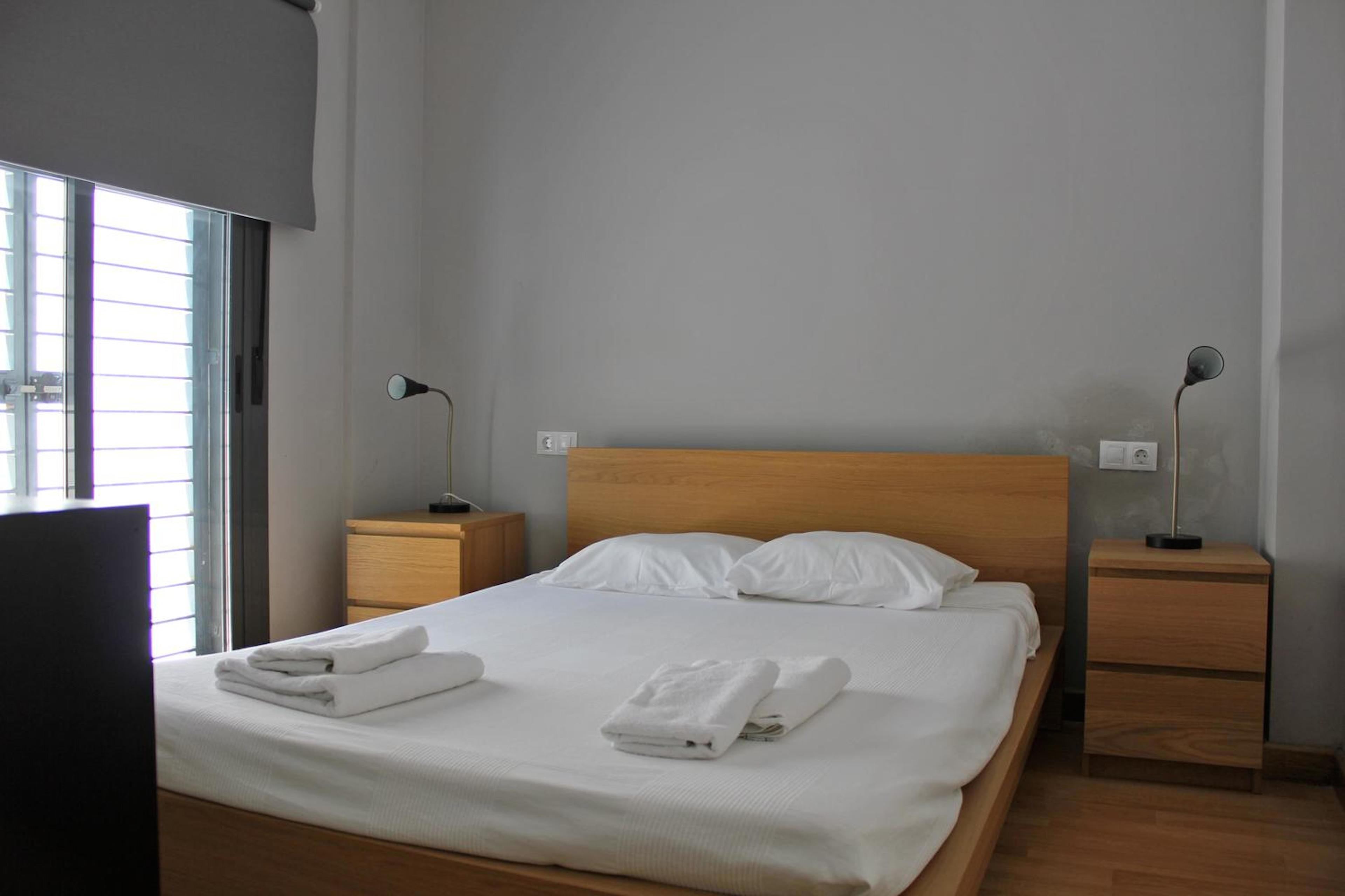 Una cama bien tendida con sábanas y toallas blancas, dos mesitas de noche con lámparas y una ventana con persianas.