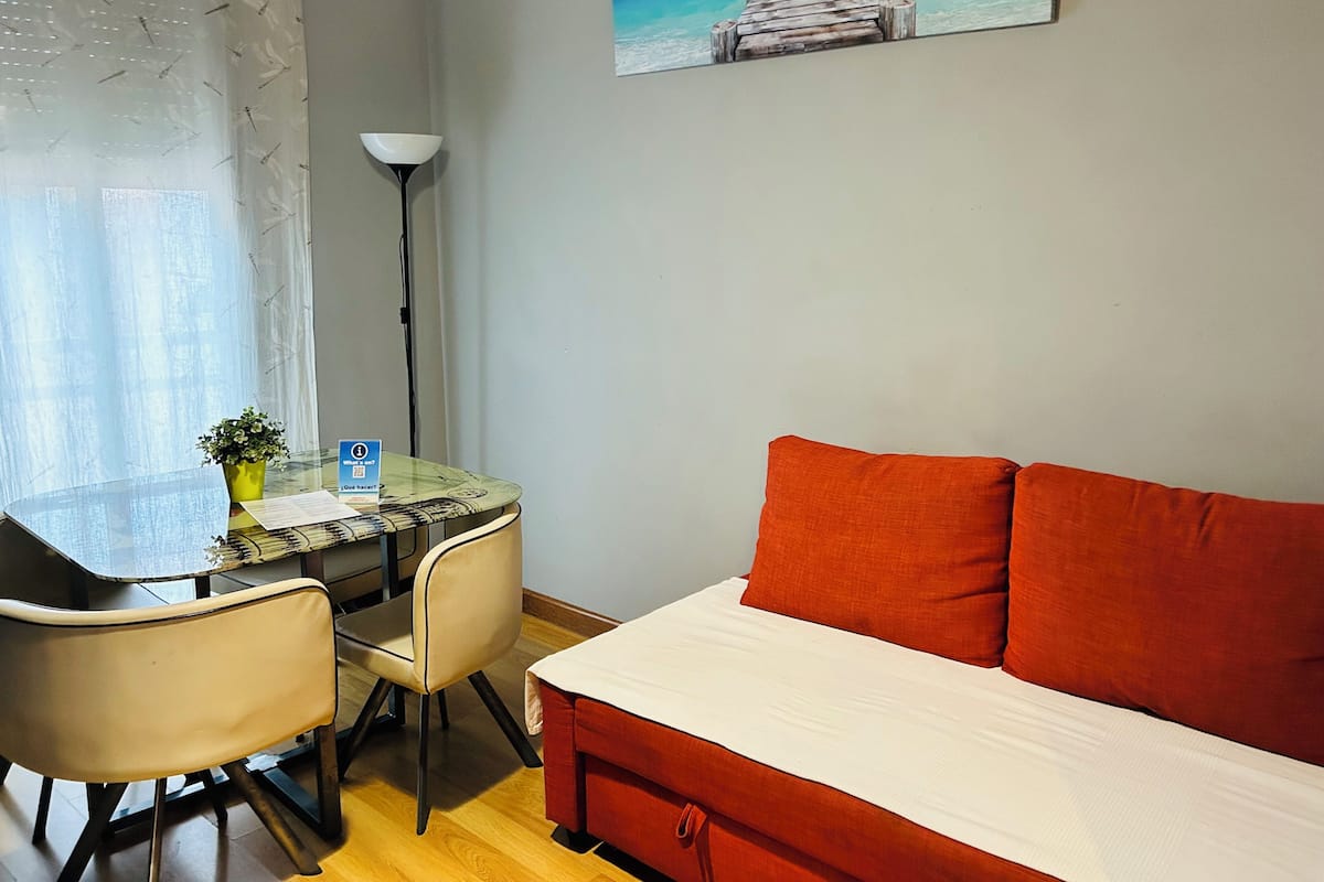 A room with a bed, a desk with a chair, a lamp, a window with sheer curtains, and a painting of a pier.