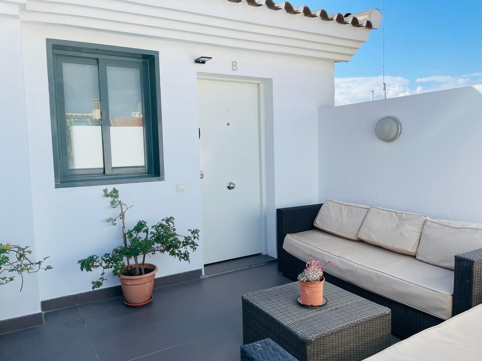 A balcony with a white wall, a door marked with the letter 'B', a window, and a beige sofa with cushions.