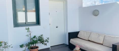 Un balcón con un muro blanco, una puerta identificada con la letra 'B', una ventana y un sofá beig con cojines.