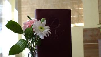 A clear glass vase with flowers on a wooden table. Behind the vase, there is a dark-colored booklet with embossed text.