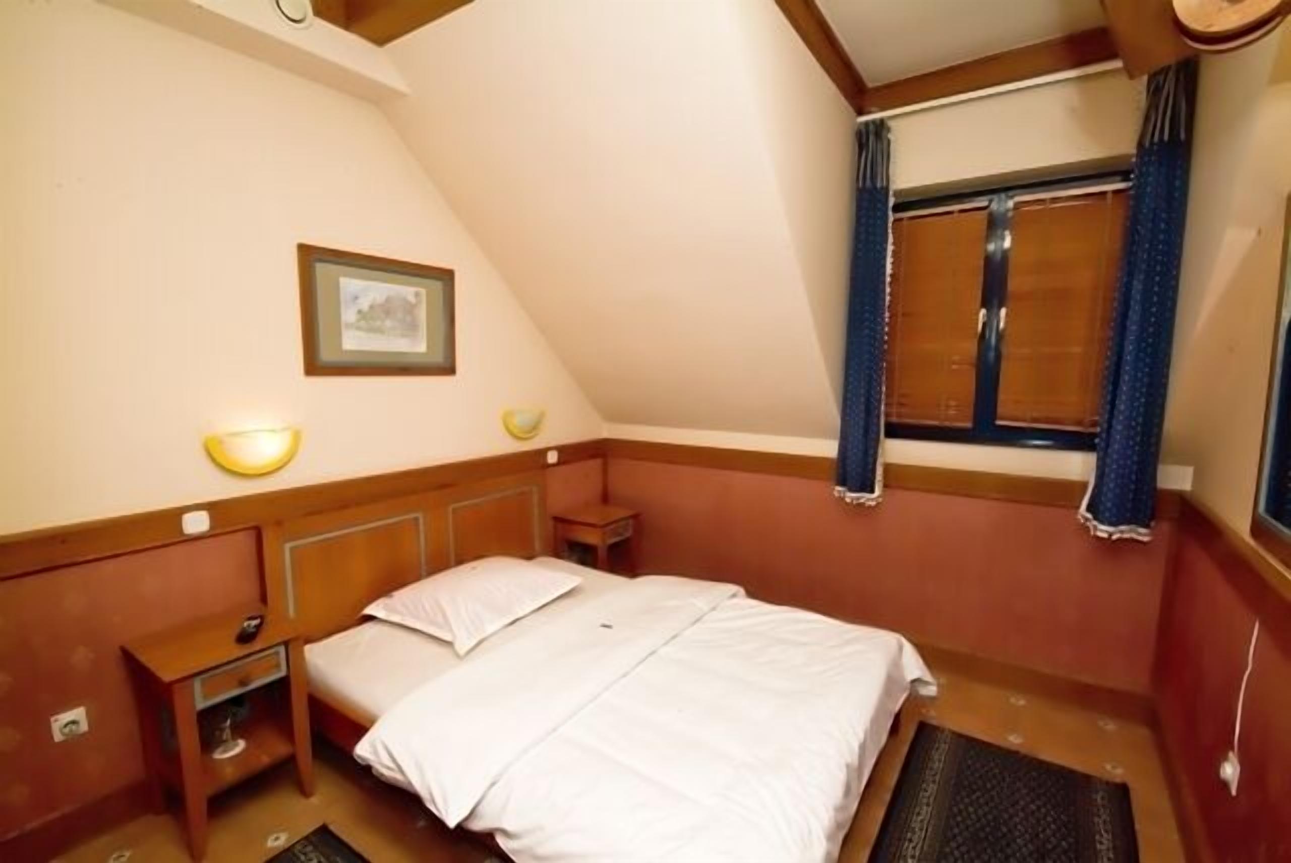 A single bed with a wooden headboard, a bedside table with a lamp, a framed picture on the wall, and a window with curtains.