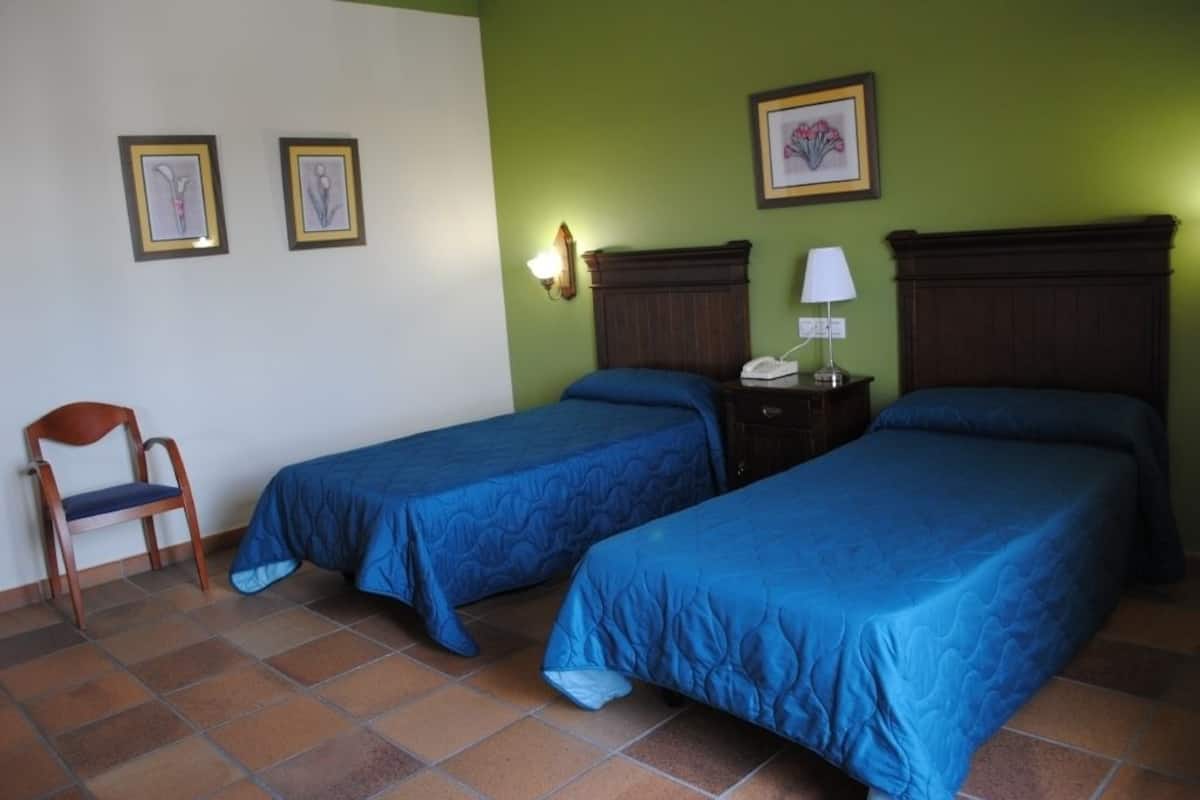 Una habitación con dos camas individuales, una silla, una mesita de noche con lámpara y cuadros en la pared.