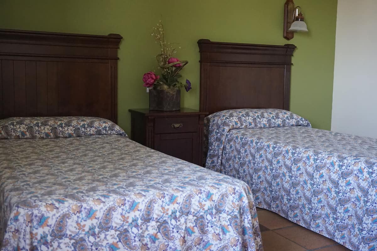 Dos camas individuales con ropa de cama floral, una mesita de noche con una planta en maceta y una lámpara de pared.