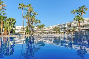 A pool with clear blue water reflecting palm trees and a hotel building with multiple balconies and sun loungers. - FERGUS Club Palmanova Park (Calvia)