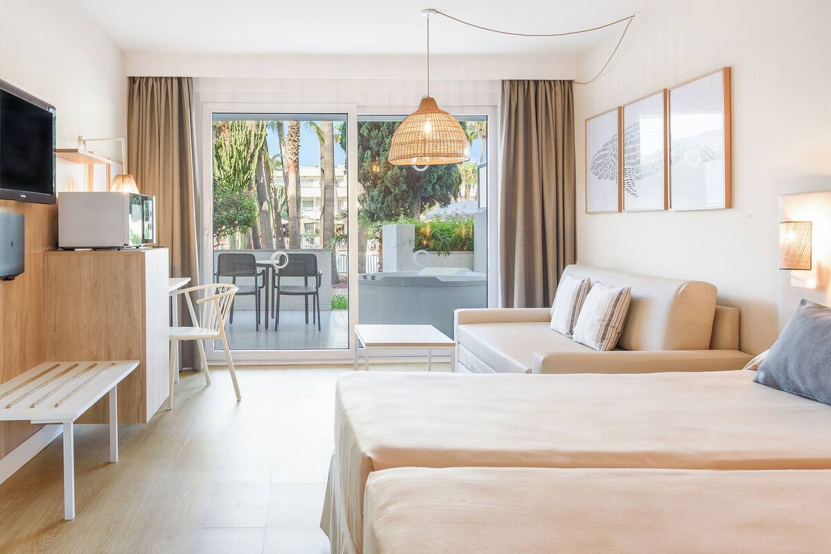 A hotel room with a bed, a desk, a chair, a TV, and a balcony with a view of palm trees and buildings.