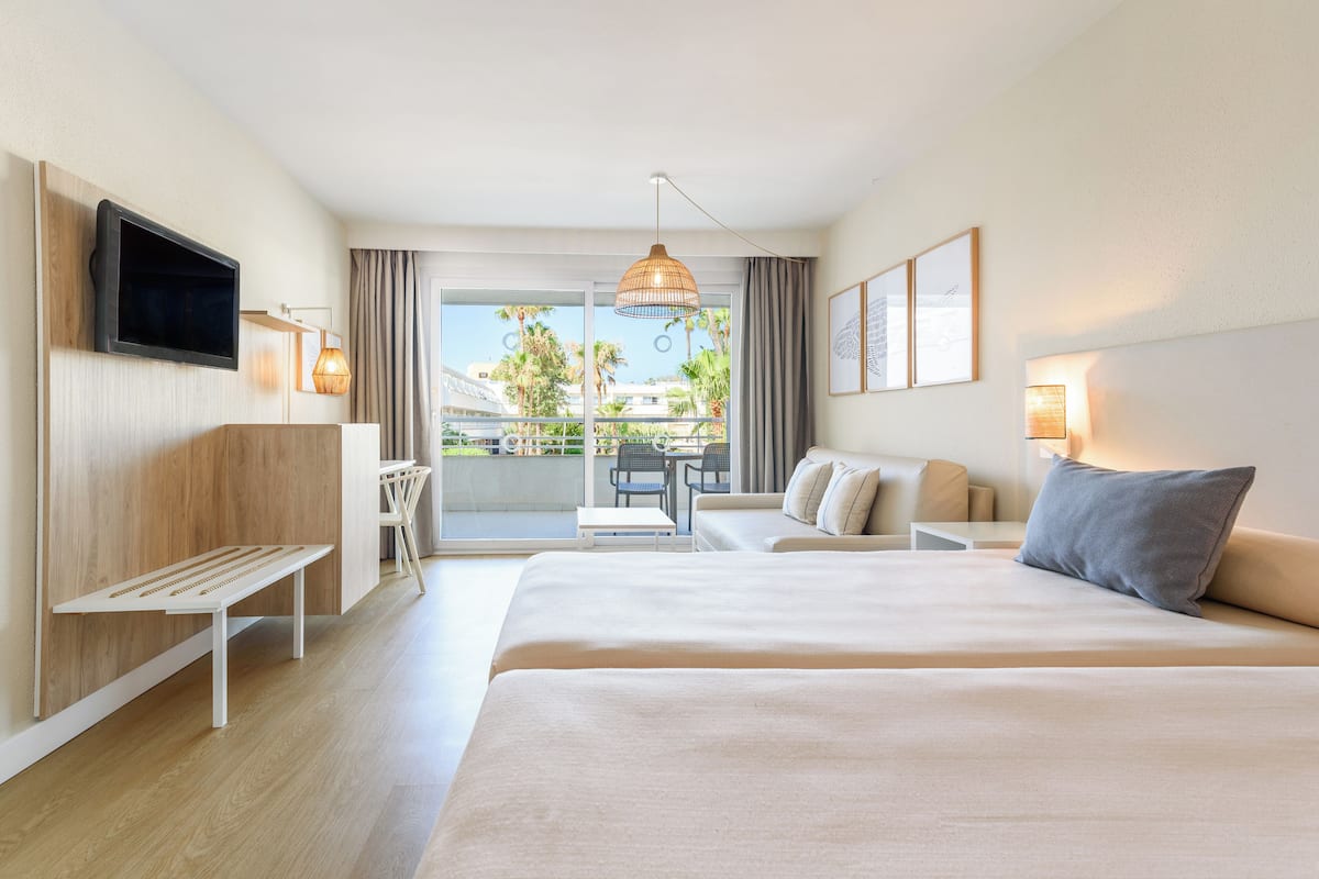 A hotel room with a large bed, a desk, a chair, a TV mounted on the wall, and a balcony with a view of palm trees and buildings.