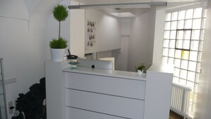 A reception desk with a potted plant, a telephone, and a small table with items. There is a window with blinds and a radiator. - Hotel Sechzehn (Leverkusen)