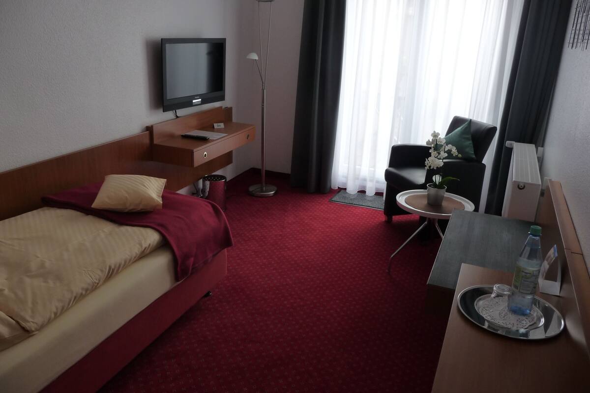 A hotel room with a bed, a desk with a chair, a TV, a floor lamp, and a window with curtains.