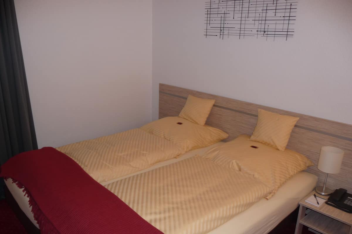 A double bed with a wooden headboard, two pillows, a red throw blanket, a bedside table with a lamp, and a wall art piece.