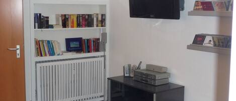 Una habitación con un radiador blanco, un televisor negro montado en la pared, una estantería con libros y una pequeña mesa con dispositivos electrónicos.