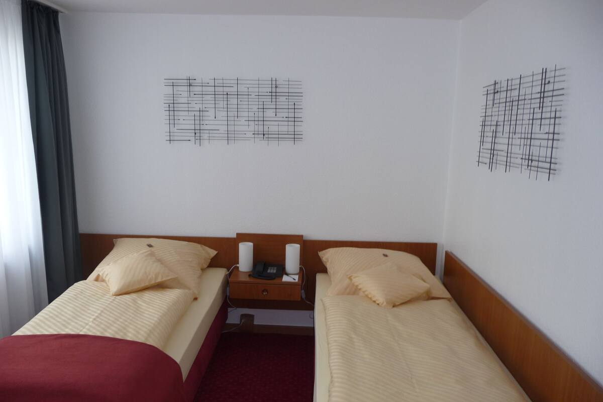 A hotel room with two single beds, a wooden nightstand with a phone and a lamp, and abstract wall art.