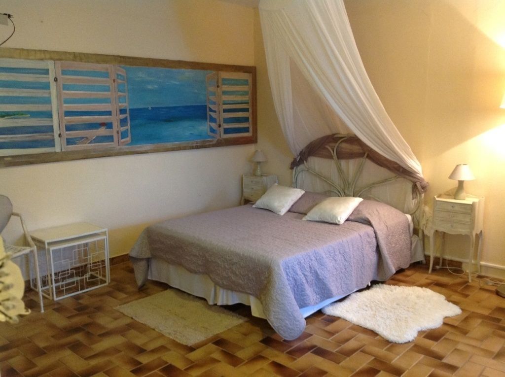 Un dormitorio con cama, mesita de noche, alfombra blanca y un cuadro de una playa. La cama tiene dosel.