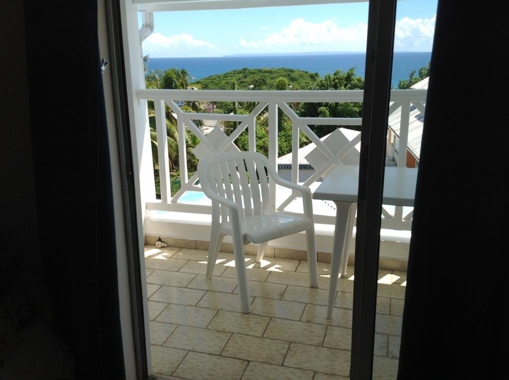 A balcony with a white plastic chair and table, overlooking a scenic view of the ocean and greenery.